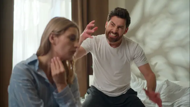 Un hombre mueve sus manos agresivamente mientras su rostro muestra enfado al parecer hacia su pareja que se encuentra sentada y pone su mano en su cuello.