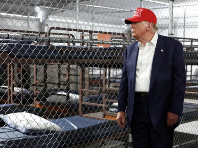 El presidente de EE. UU. Donald Trump con gorra roja de MAGA posa frente a una verja con literas detrás, durante su visita al centro temporal de detención de migrantes conocido de manera informal como 