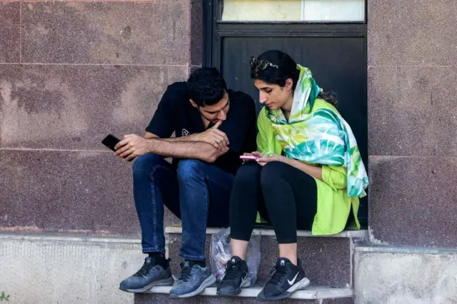 یک دختر بدون حجاب در کناری پسری نشسته و هر دو به گوشی موبایلی که دست دختر است نگاه میکنند