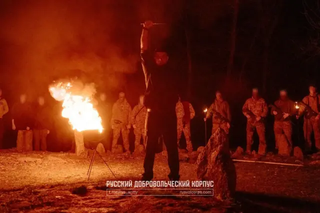 Нічна сцена, освітлена сильним полум’ям від вогнища чи факела на металевій підставці. Попереду стоїть чоловік у темному одязі, піднявши руку з довгим предметом (ймовірно, ножем чи ритуальним клинком). Позаду в ряд вишикувано групу людей у камуфляжній формі, обличчя яких розмиті. Дехто тримає факели. Місцевість схожа на лісову галявину, навколо видно дерева та камені.