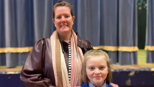 Terri Lamberton está de pie junto a su hija, Gail. Tiene el cabello castaño recogido. Lleva una chaqueta de cuero burdeos sobre una blusa a rayas blancas y negras, y una bufanda multicolor. Gail tiene el pelo largo y rubio y viste su uniforme escolar.