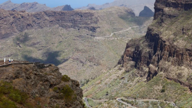 A view of a large cliff face looking down on to a winding road below in the Masca area of northern Tenerife. The land is dotted with cacti and the terrain is rough and hilly.