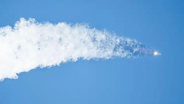 Космическая ракета взлетает в чистое голубое небо, оставляя за собой огромный серый след, который постепенно расширяется.