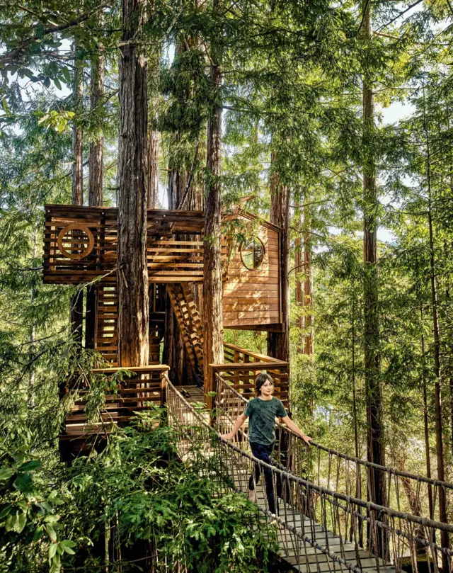 Una casa en un árbol de madera, de dos pisos, que está conectada a un puente colgante por el cual camina una mujer. 