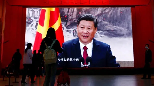 Şi Jinping dev ekranda Çin bayrağı yanında görülüyor, arkası dönük insanlar ekran önünden geçiyor