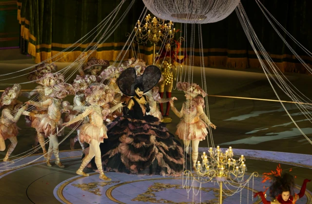 Opera na zatvaranju olimpijskih igara, žena u dugoj, bogatoj haljinisa velikim crnim šeširom na glavi, okružena balerinama