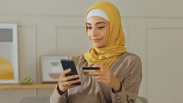 Una mujer musulmana mira un teléfono inteligente mientras sostiene una tarjeta bancaria en su mano.