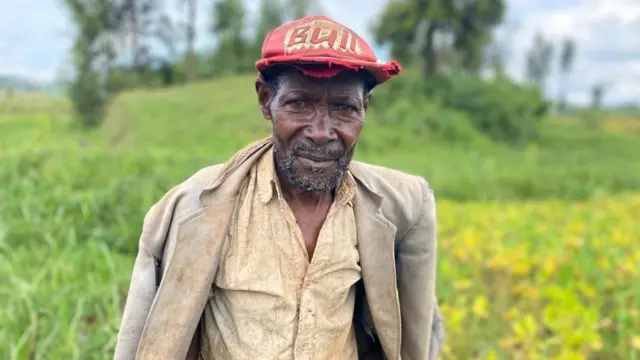 Umugabo wambaye ikoti, ishati n'ingofero itukura ahagaze mu murima urimo imyaka