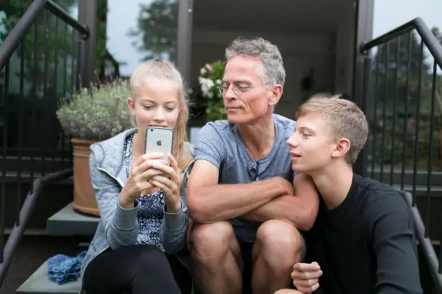 Pai ao lado de dois filhos. Uma menina está com celular na mão e ele observa