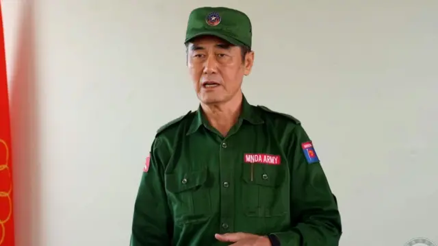 စစ်စိမ်းရောင်ဝတ်စုံကိုဝတ်ဆင်ထားပြီး မတ်တပ်ရပ်အနေအထားနဲ့စကားပြောနေတဲ့ ကိုးကန့်တပ်ခေါင်းဆောင် ဖုန်တာရွှင် ကိုတွေ့ရပါတယ်။