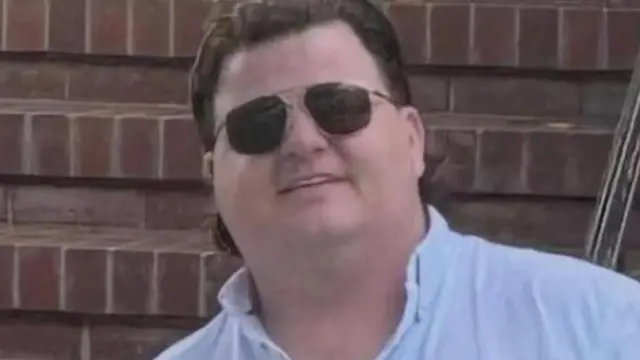 Thomas Casey tiene cabello castaño, lleva gafas de sol negras y una camisa azul pálido. Sonríe frente a una pared de ladrillos.