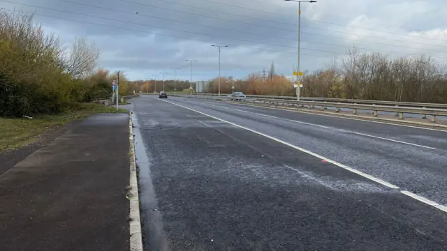La A6, con cuatro carriles, tiene dos coches. Hay árboles a ambos lados y semáforos en el centro. Hay líneas blancas en la carretera.