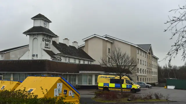 تصویر یکی از هوتلهای محل اقامت پناهندهگان. در مقابل هوتل یک موتر نوع امبولانس پولیس ایستاده است. هوتل دست کم چهار طبقه دارد.