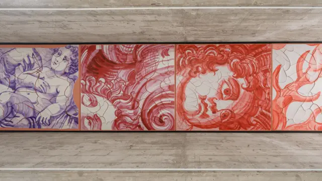 Um painel horizontal reúne desenhos em tons de vermelho e azul que remetem a figuras humanas, rostos e formas orgânicas em movimento. A obra, instalada sob uma estrutura de concreto aparente, mistura referências aos azulejos tradicionais com traços fluidos e fragmentados.