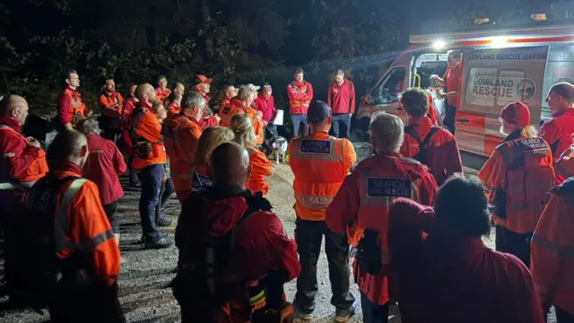 Un grupo de voluntarios, vestidos con ropa de color naranja y rojo, se reúnen alrededor de una camioneta iluminada, como si estuvieran recibiendo instrucciones.