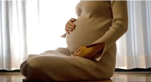 ifoto y'umugore utwite wicaye hasi afashe ku nda ye, yambaye imyenda iri mu ibara rya kaki