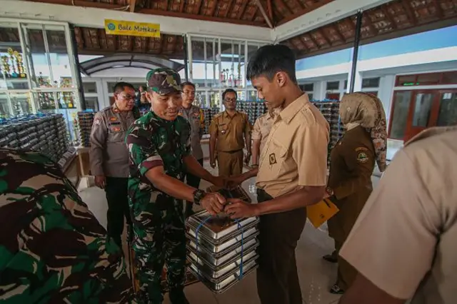 Seorang tentara Angkatan Darat Nasional Indonesia membagikan wadah makanan bergizi gratis (MBG) kepada seorang siswa di Sekolah Menengah Atas Negeri 20 di Bandung, Jawa Barat, Indonesia, pada tanggal 14 April 2025. 