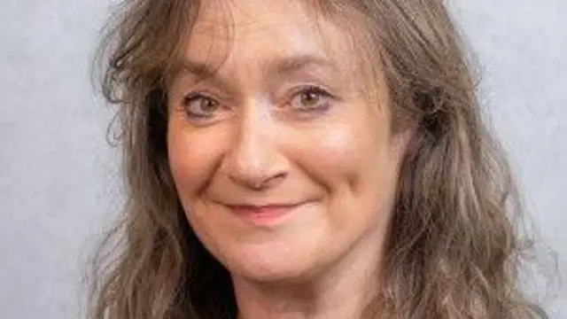 Retrato de Daphne Caine sobre un fondo gris. Tiene una sonrisa tenue y hoyuelos, y cabello castaño y gris.