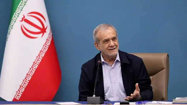 Koyu mavi bir ceket ve açık mavi yakalı bir gömlek giyen ve büyük bir İran bayrağının sağında bir masada oturan İran Cumhurbaşkanı Mesud Peşkiyan, 24 Şubat 2026'da bir konuşma yapıyor.