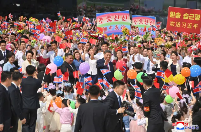 Chiński przywódca Xi Jinping wita zebrany tłum podczas wizyty w Korei Północnej. Za nim stoi jego gospodarz, Kim Dzong Un. 