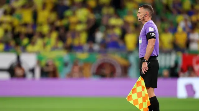 Un hombre con camiseta morada, pantalón negro y auriculares está de pie en la línea de banda, sosteniendo una bandera a cuadros naranja y amarilla a su lado. Mira al otro lado del campo.