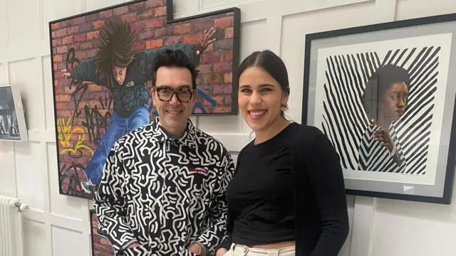 Two artists, one male and one female, stand in front of their artwork in an exhibition hall. One piece of artwork depicts a young person jumping in front of a graffitied brick wall. Another depicts a black lady staring ahead with a striped black and white top in front of similarly striped black and white background.