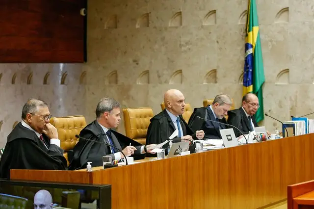 Dino, Mendonça, Moraes, Toffoli e Gilmar Mendes sentados lado a lado no plenário do STF, ventindo terno e suas togas de ministro; Moraes fala ao microfone enquanto os demais escutam
