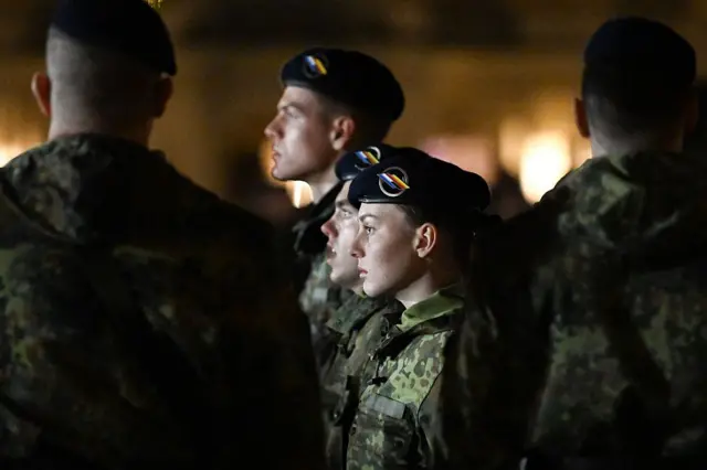 Bir kadın asker oldukça ciddi şekilde duruyor, yanında omuzları görünen askerler var.