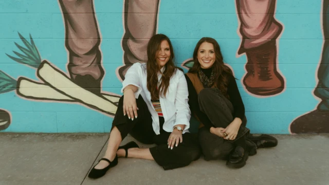 Jacqueline and Alexa are sitting on the ground in a professional photo in front of a cartoon-style mural on a wall, showing two mens' legs and two large carrots. They are both smiling to camera in a posed shot.