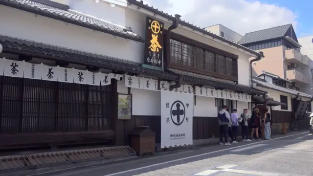 道路に面した和風の建物。白壁に茶色の格子、かわらのひさしや屋根。そこに、丸に十字の紋や「中村銘茶」「茶」といった文字が書かれた、白いのれんや幕がかかっている。ひさしの上には茶色い板に金で「茶」と書かれた看板も立つ。店の前に数人の行列ができている