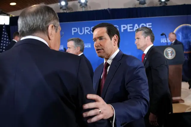 Lavrov está de espaldas a la cámara; Rubio le dice algo y le toca el hombro con la mano. El secretario de Defensa de Estados Unidos, Pete Hegseth, está detrás de ellos, frente a un cartel que dice "En busca de la paz", en Anchorage, Alaska, el 15 de agosto de 2025.