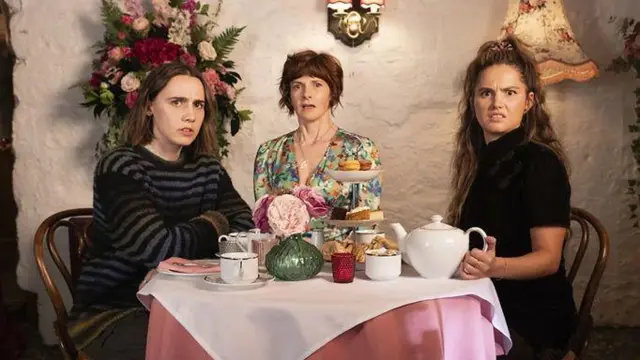 Кадр із серіала "Ці хоробрі дівчатка". Три жінки сидять за столом з чаєм і тістечками, дивлячись у камеру одночасно з подивом і легким роздратуванням.
