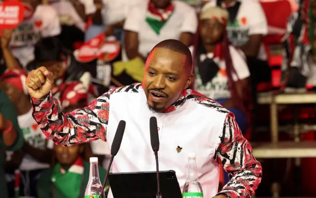 Boniface Mwangi arimo kuvuga ijambo kuri za microphones yambaye ishati y'umweru n'amabara y'umutuku n'umukara