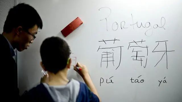 Aluno copia os ideogramas da palavra "Portugal" em mandarim em um quadro branco, sob a supervisão do professor