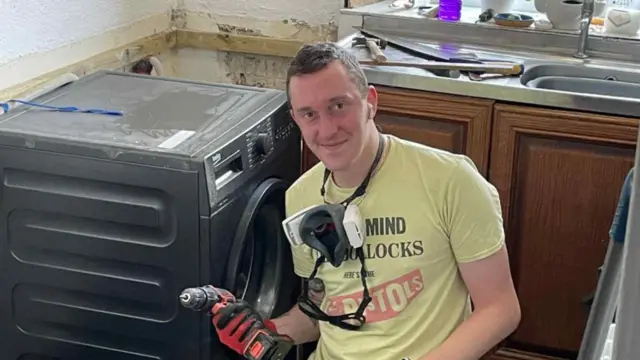 Um homem está sentado ao lado de uma máquina de lavar, segurando uma furadeira enquanto realiza reparos. Ele veste uma camisa da banda Sex Pistols.