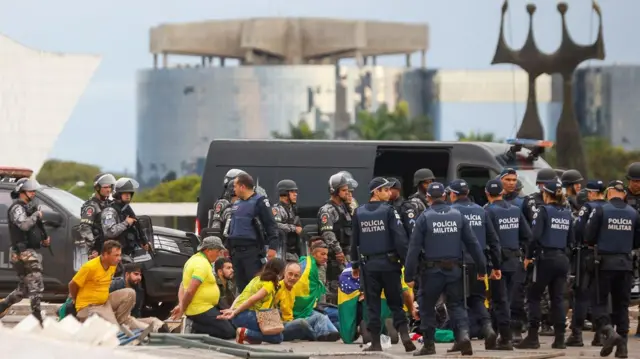 Pessoasmines onabetcamisa verde e amarela sentadas com as mãos algemadas; eles estão rodeados por policiais na Praça dos Três Poderes