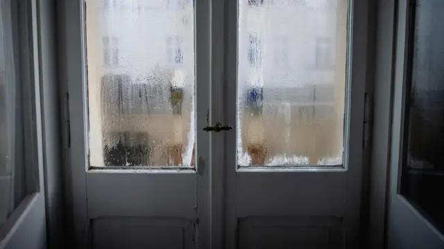 Zamarznięte okno w starym berlińskim mieszkaniu widziane od środka; po szybach spływa skroplona para. Z zewnątrz wpada dzienne światło, a przez mglistą, zamarzniętą powierzchnię widać zarys innego budynku mieszkalnego.