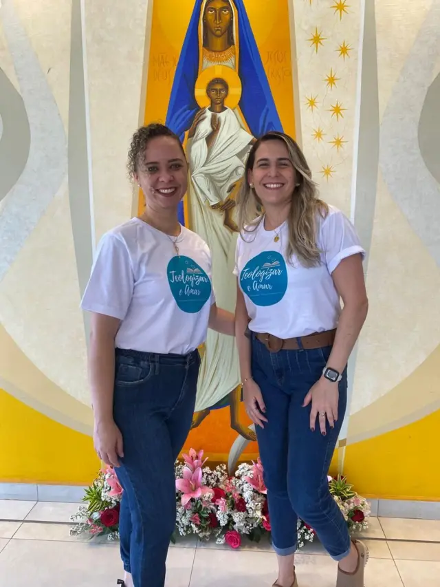 A bancária Pamella Barbosa (cabelos encaracolados) e sua noiva, em eventos da igreja - casal de duas mulheres com camisetas brancas e calças jeans azuis, na frente de uma imagem religiosa e flores
