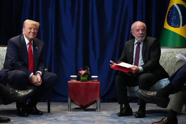 presidentes Luiz Inácio Lula da Silva e Donald Trump apertando as mãos durante encontro na Malásia, no domingo, 26 de outubro. Ambos sorriem, com bandeira do Brasil ao fundo