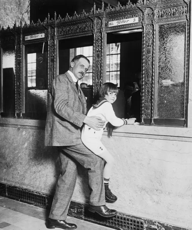 Amadeo Giannini cargando al actor infantil Jackie Coogan para que alcance la ventanilla del cajero.