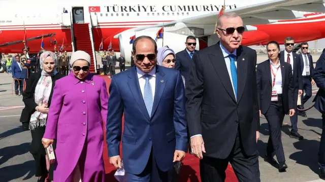 Erdoğan ve Sisi, güneş gözlüklü geride pembe padesülü bir kadın görünüyor. Liderlerin ortasından Emin Erdoğan'ın başı seçilebiliyor. Onları protokol izliyor. Tümü kırmızı halıda, geride Türkiye Cumhuriyeti yazılı bir jet var.