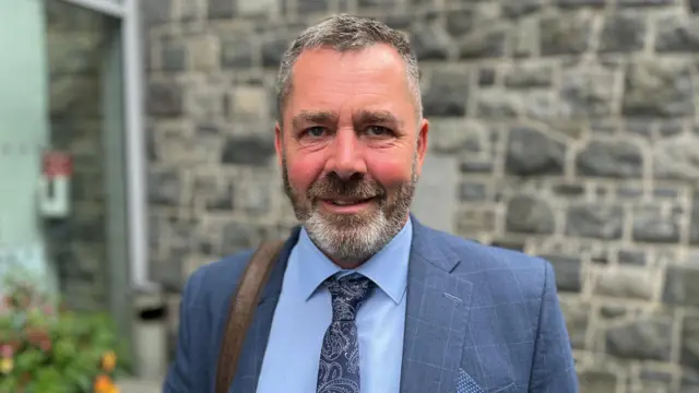 Diputado Marc Leadbeater: Un hombre de cabello negro y canoso, con una barba similar, mira a la cámara. Lleva un traje con estampado de rayas azules, una camisa azul abotonada y una corbata azul estampada.