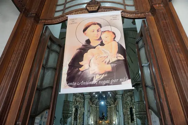  "Santo Antônio, rogai por nós". Ele está afixado no que parece ser o batente de uma porta e é possível ver o altar ornamentado de uma igreja ao fundo