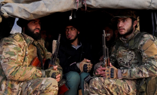 Elinde uzun namlulu silah olan iki uniformalı bir askeri aracın arkasında oturuyor. İkisi de farklı noktalara bakıyor. 