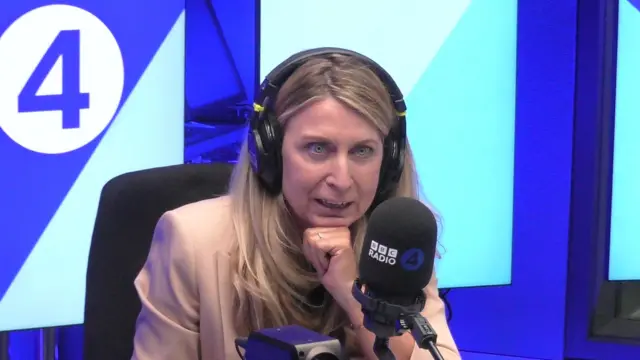 明るい色の長髪で、ピンクのスーツを着た女性が、ラジオのマイクの前で話している