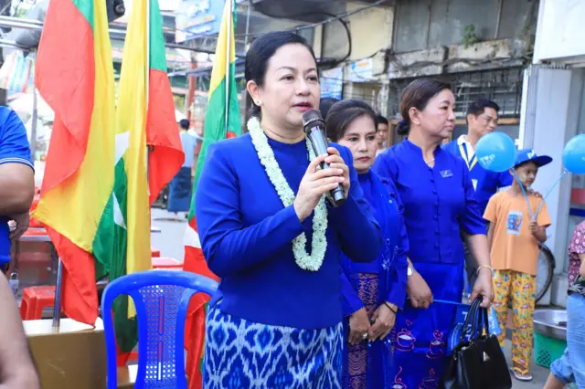 အပြာရောင်မြန်မာရိုးရာဝတ်စုံ၊ စံပယ်ပန်းကုံးဆွဲထားတဲ့ ဝမ်းဆက်နဲ့ ဒေါ်သက်သက်ခိုင်၊ သူ့အနောက်မှာရပ်နေတဲ့ ပါတီဝင်အမျိုးသမီး ၂ ဦး၊ မြန်မာနိုင်ငံတော်အလံ 