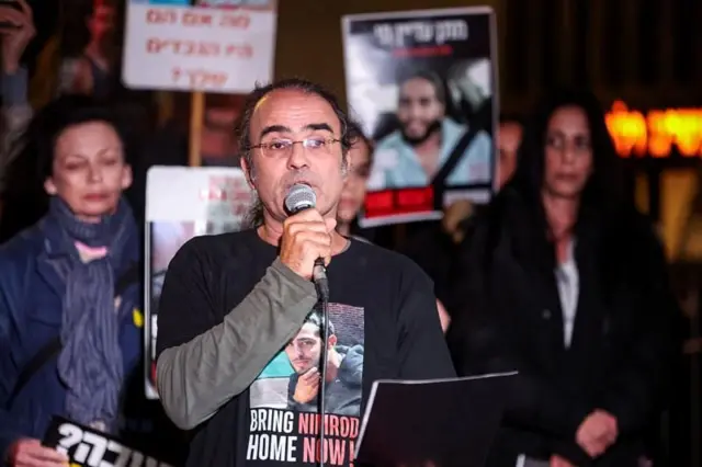 Homem falando em manifestação, com pessoas atrás segurando cartazes