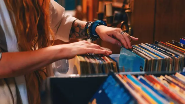 Braços de uma mulher verificando uma caixa de discos de vinil com capas coloridas em uma loja de discos