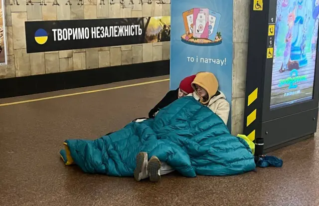 Люди у куртках під спальником сплять на підлозі у метро. На стіні - плакат 