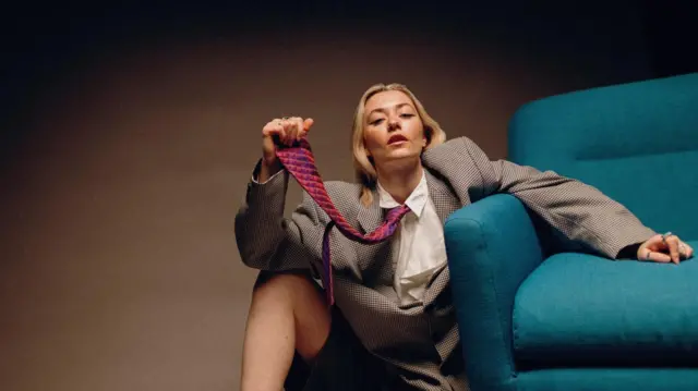 Una mujer rubia con una chaqueta deportiva, corbata roja y azul y camisa blanca está sentada en el suelo junto a una silla azul.